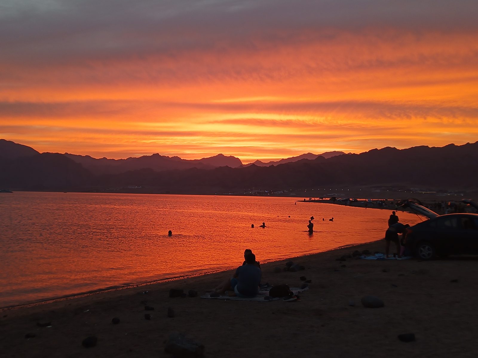 Orange glühender Abendhimmel über dem Meer, dahinter Bergketten, am Strand kleine Silhouetten von Badenden