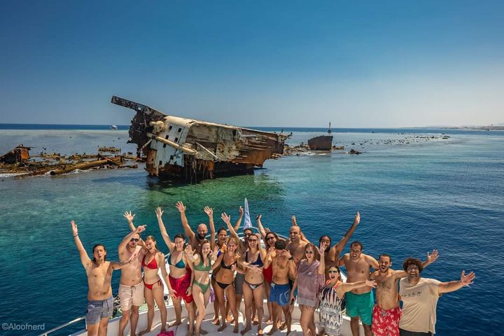 Eine fröhliche Gruppe in Badekleidung auf einem Boot, dahinter ein gestrandetes Schiffswrack auf einem Riff